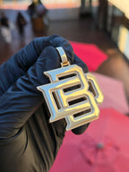Interlocked BD gold monogram pendant held at an angle in warm light, showing the matte beveled edge and squared 7mm tube bail